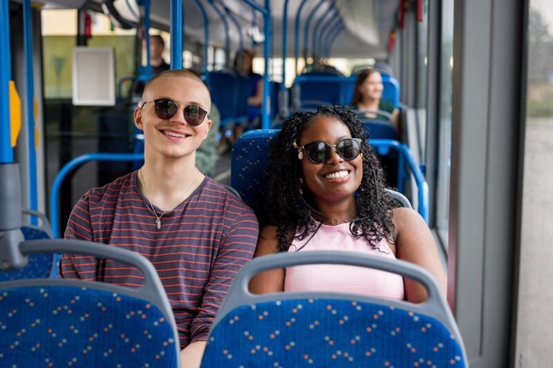 Tyttö ja poika istuvat aurinkoisesti hymyille bussin kyydissä aurinkolasit päässä.
